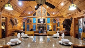 A log cabin interior with a stone fireplace, wooden walls, and a granite dining table set for four. - Deer Ridge Cabin - Hochatown - Hot Tub - Firepit - Spacious Deck - Large Yard (Broken Bow)