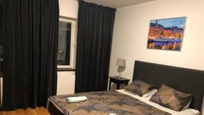 Ein Hotelzimmer mit Bett, Fenster mit Vorhängen, Nachttisch mit Lampe und einem Bild an der Wand.