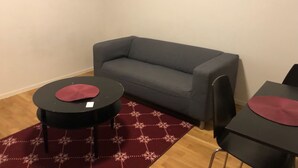 Ein kleiner Wohnbereich mit einem grauen Sofa, zwei schwarzen Tischen mit roten Kissen und einem gemusterten roten Teppich auf Holzdielen.