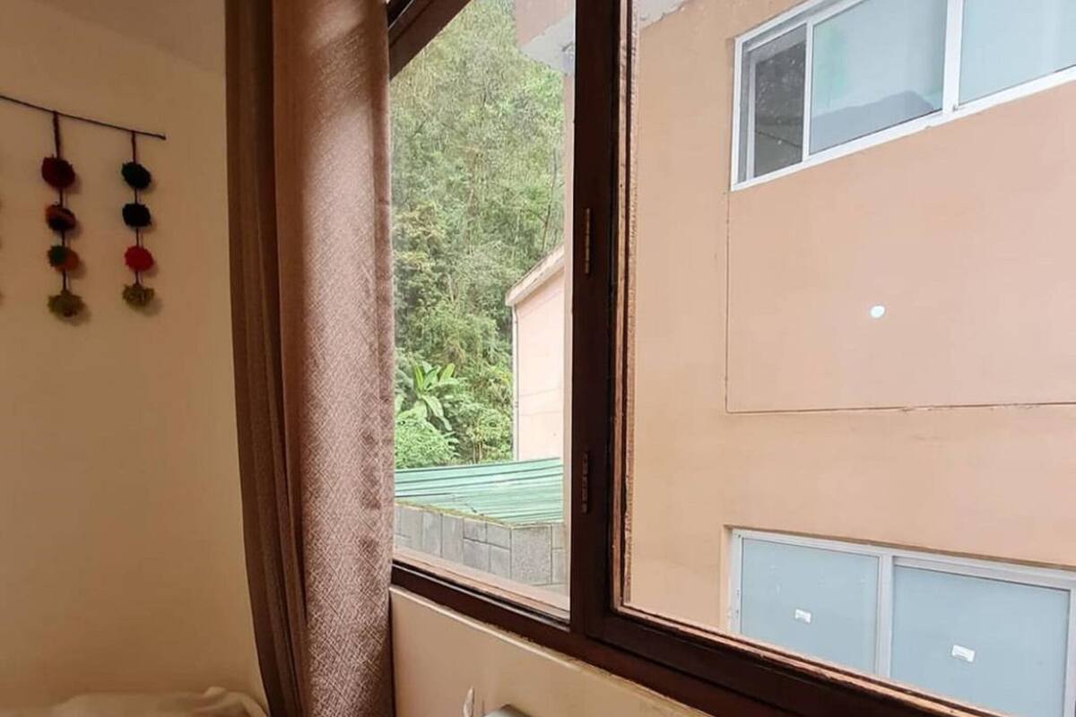 A bedroom with a bed, a window with a view of a building and greenery, and a wall-mounted storage unit.