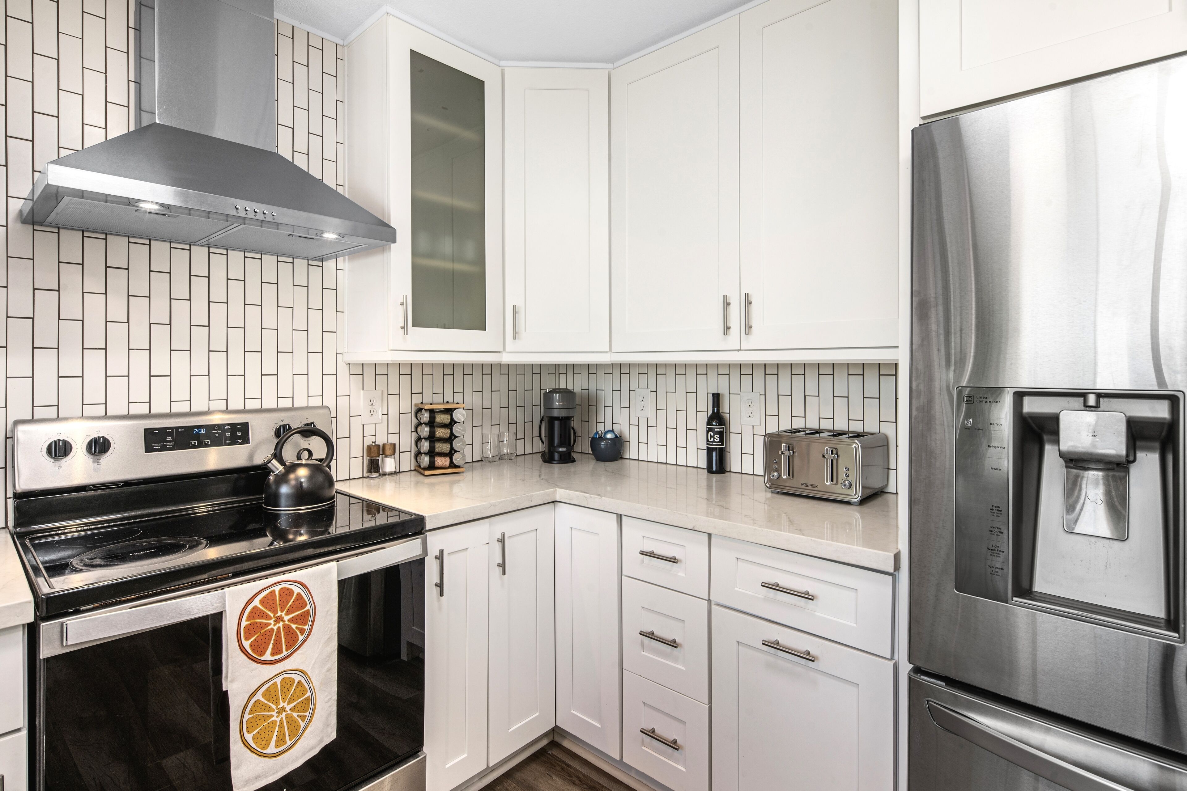 A modern kitchen with white cabinets, a stainless steel range hood, a built-in oven, a refrigerator, and a coffee maker.