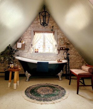 A bathroom with a freestanding bathtub, a wooden chair, a patterned rug, and a stone wall.