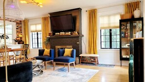 A living room with a fireplace, two blue armchairs, a black sofa, a wooden coffee table, and a television above the fireplace. - Vintage Charm — 5-Min Walk to Downtown & Campus (Eugene)