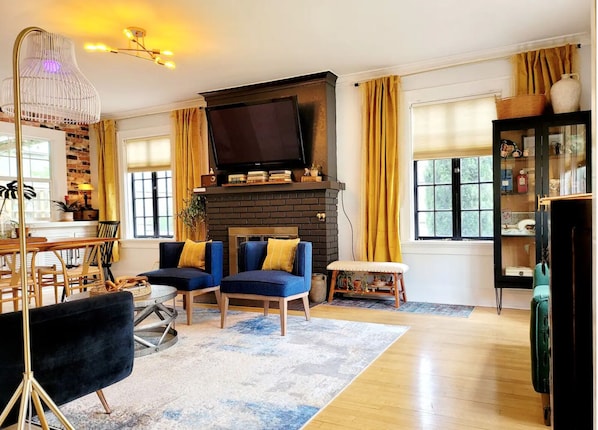 A living room with a fireplace, two blue armchairs, a black sofa, a wooden coffee table, and a television above the fireplace.