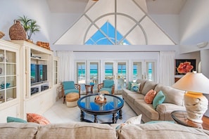 A spacious living room with a glass coffee table, a sofa, and a TV. There are multiple windows offering a view of the ocean.