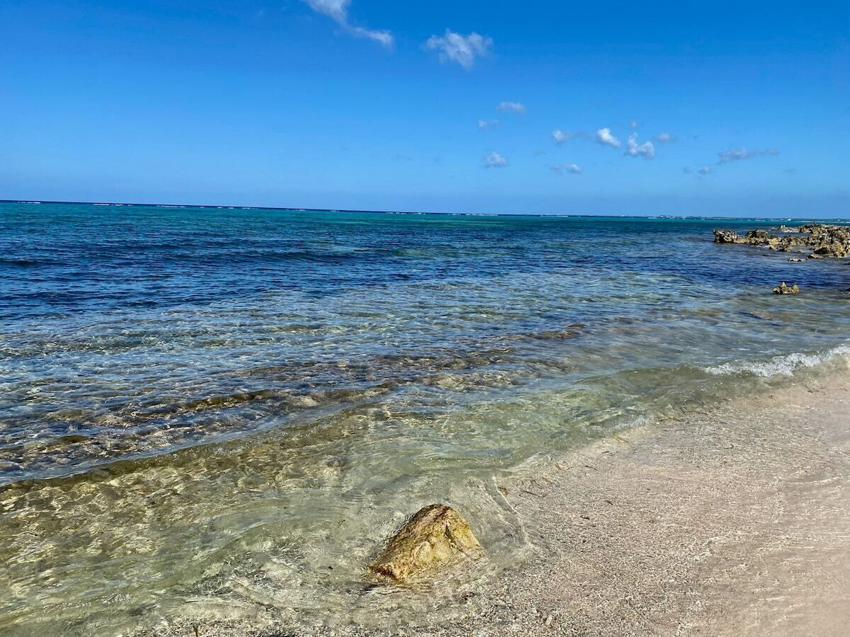 A beach with clear shallow waters, a visible rock in the foreground, and a distant rocky shoreline.