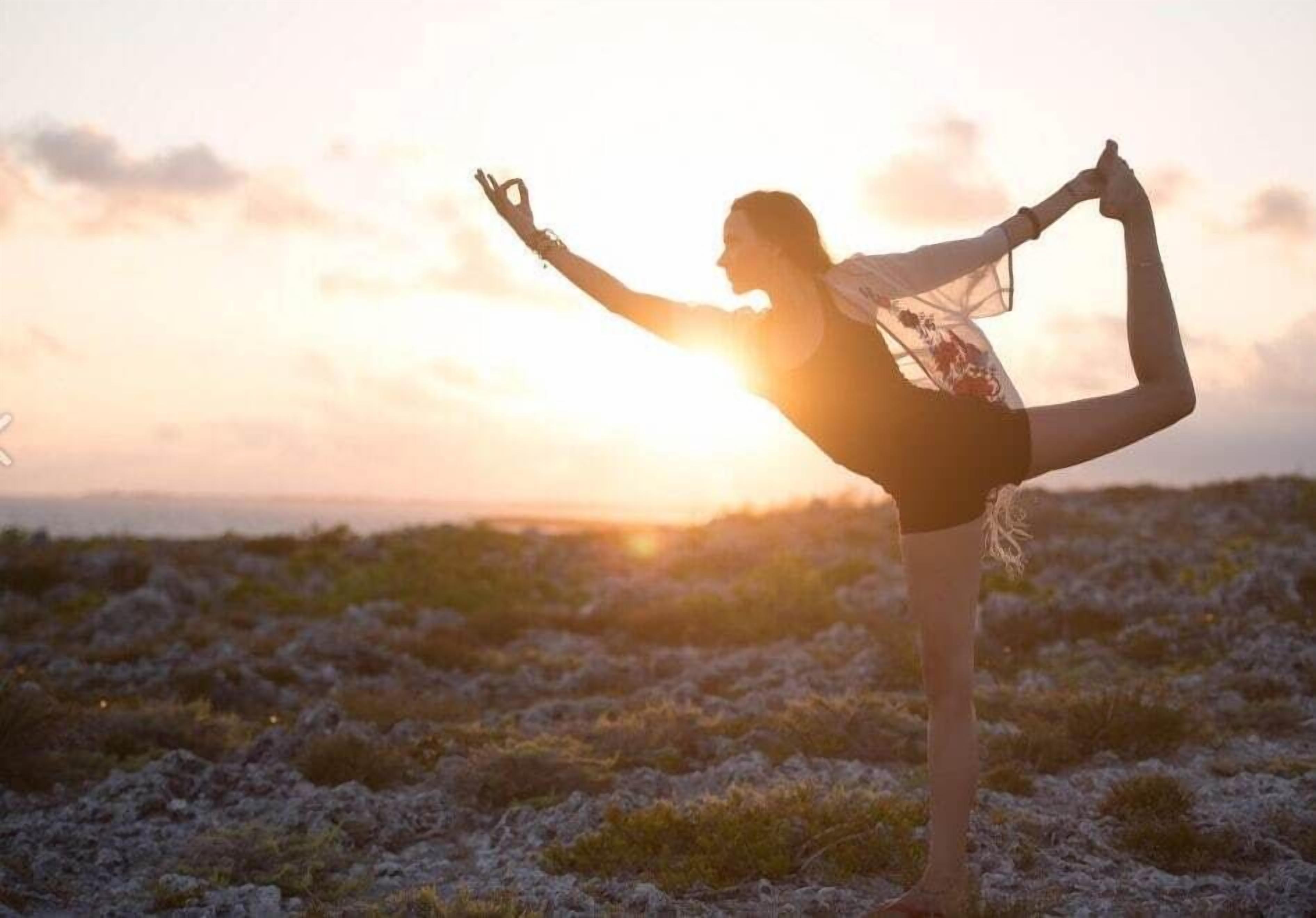 En person utför en yogaposition på en gräsklädd äng med en solnedgång i bakgrunden.
