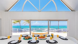 Une table à manger dressée pour quatre personnes, avec assiettes, couverts et boissons, donnant sur une piscine et l’océan.