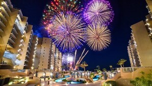 A hotel with multiple floors, outdoor swimming areas, and a view of fireworks in the background.