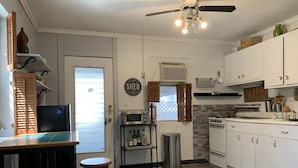 A kitchen with a refrigerator, stove, microwave, and sink. There is a window with blinds, a ceiling fan, and a trash can.