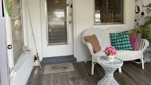 A white door with a metal grill, a wicker chair with patterned cushions, and a small white table with a vase of flowers.