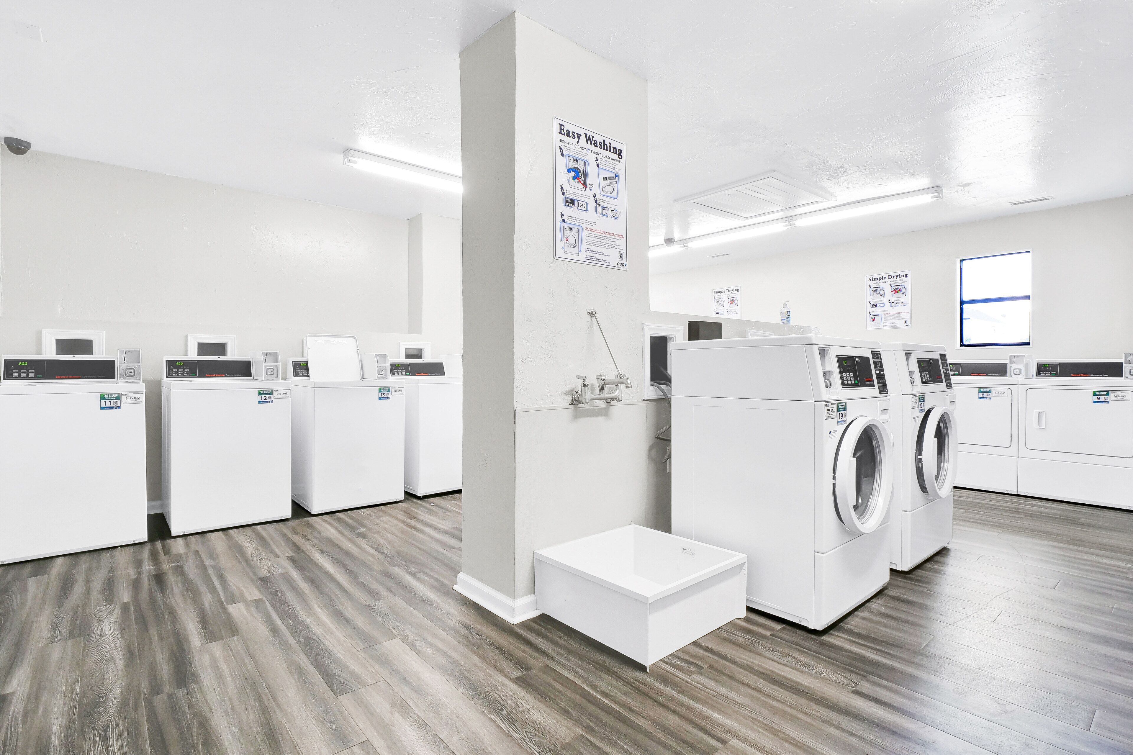 A clean, modern laundry facility with multiple washing machines and dryers, a step for loading, and instructional posters on the walls.