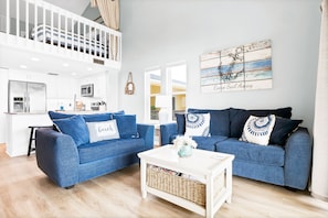 A modern living room with two blue sofas, a white coffee table, and a kitchen area in the background.