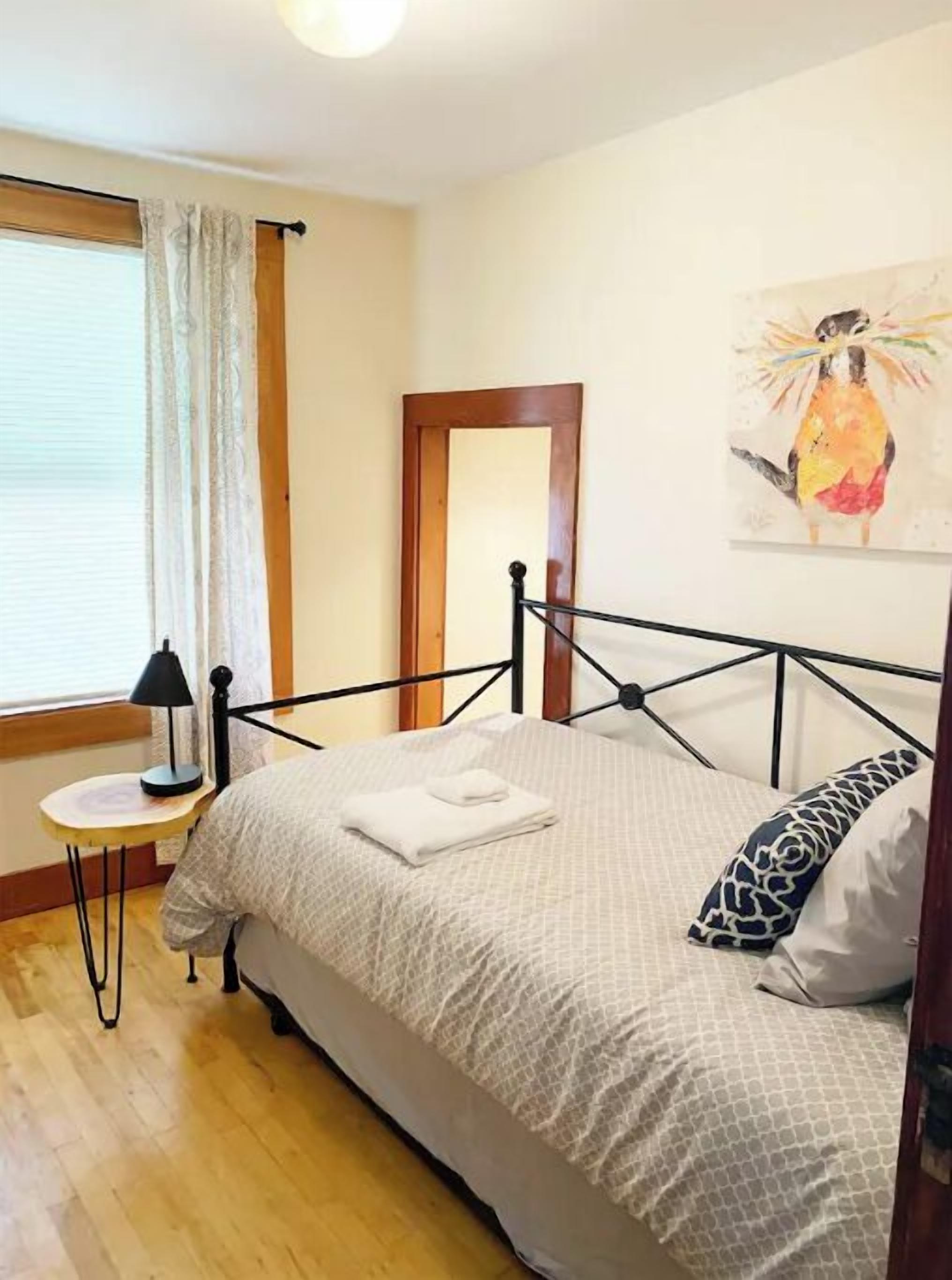 Un dormitorio con cama, mesita de noche, lámpara, ventana con cortinas y un cuadro en la pared.