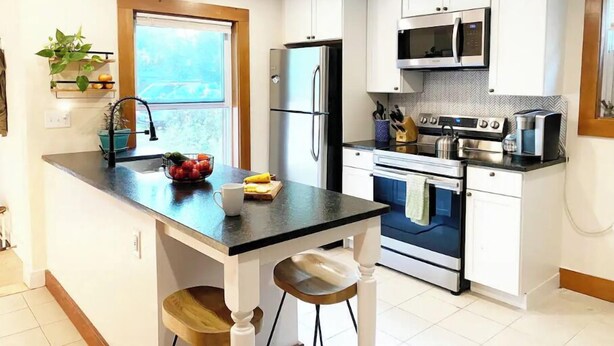 A modern kitchen with white cabinets, a stainless steel refrigerator, a microwave, and an oven. There is a window providing natural light and a small plant on the countertop.