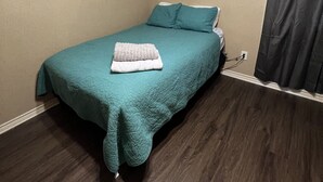 Una habitación pequeña y ordenada, con una cama, una lámpara fijada a la pared y una cortina oscura.