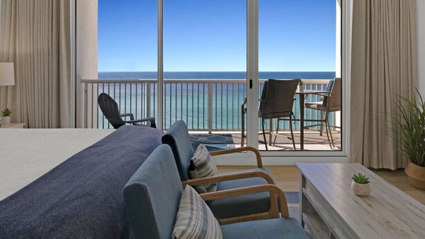 Una habitación de hotel con una cama grande, un balcón con vista al océano y una zona de estar con sillas y una mesa.