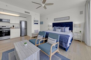 A modern hotel room with a bed, a desk, a chair, a small table, and a kitchenette.