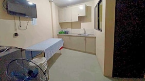 A small, compact room with a bed, a table, a TV mounted on the wall, a sink, and cabinets. - 2020 Travelers Inn (Dumaguete)