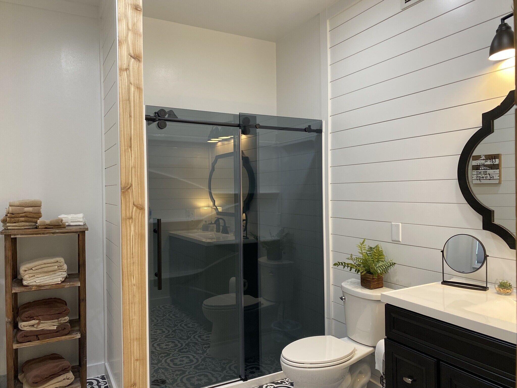 A bathroom with a glass-enclosed shower, a toilet, a sink with a mirror, and a shelf with towels.