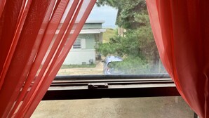 A window with red curtains partially drawn, revealing a view of a house and a parked car outside. - Beach Side Studio #5 -  Pet Friendly, WiFi, Off-Street Parking, Large Smart TV (Norfolk)