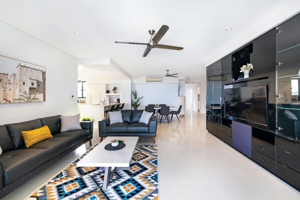 Un salon moderne avec un canapé noir, une table basse et un tapis à motifs. On aperçoit à l’arrière-plan un coin repas avec des chaises et une table.