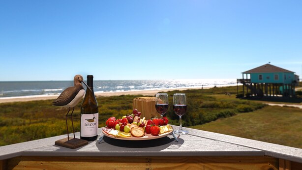Uma mesa com uma garrafa de Decoy Pinot Noir, um prato de frutas e duas taças de vinho, com vista para uma praia e uma casa sobre palafitas.