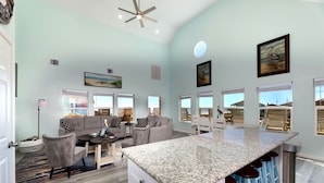 Uma sala de estar espaçosa com um sofá grande, uma mesa de centro e uma mesa de jantar. Há uma área de cozinha com bancada de granito e banquetas. O espaço possui grandes janelas com vista para o exterior.