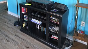 Een woonkamer met een wandtelevisie, een zwarte entertainmentkast met boeken, een lamp en een klein houten plankje.