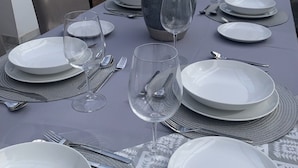 Una mesa de comedor dispuesta con platos blancos, vasos y cubiertos, un florero con flores y vistas al exterior a través de ventanas grandes.