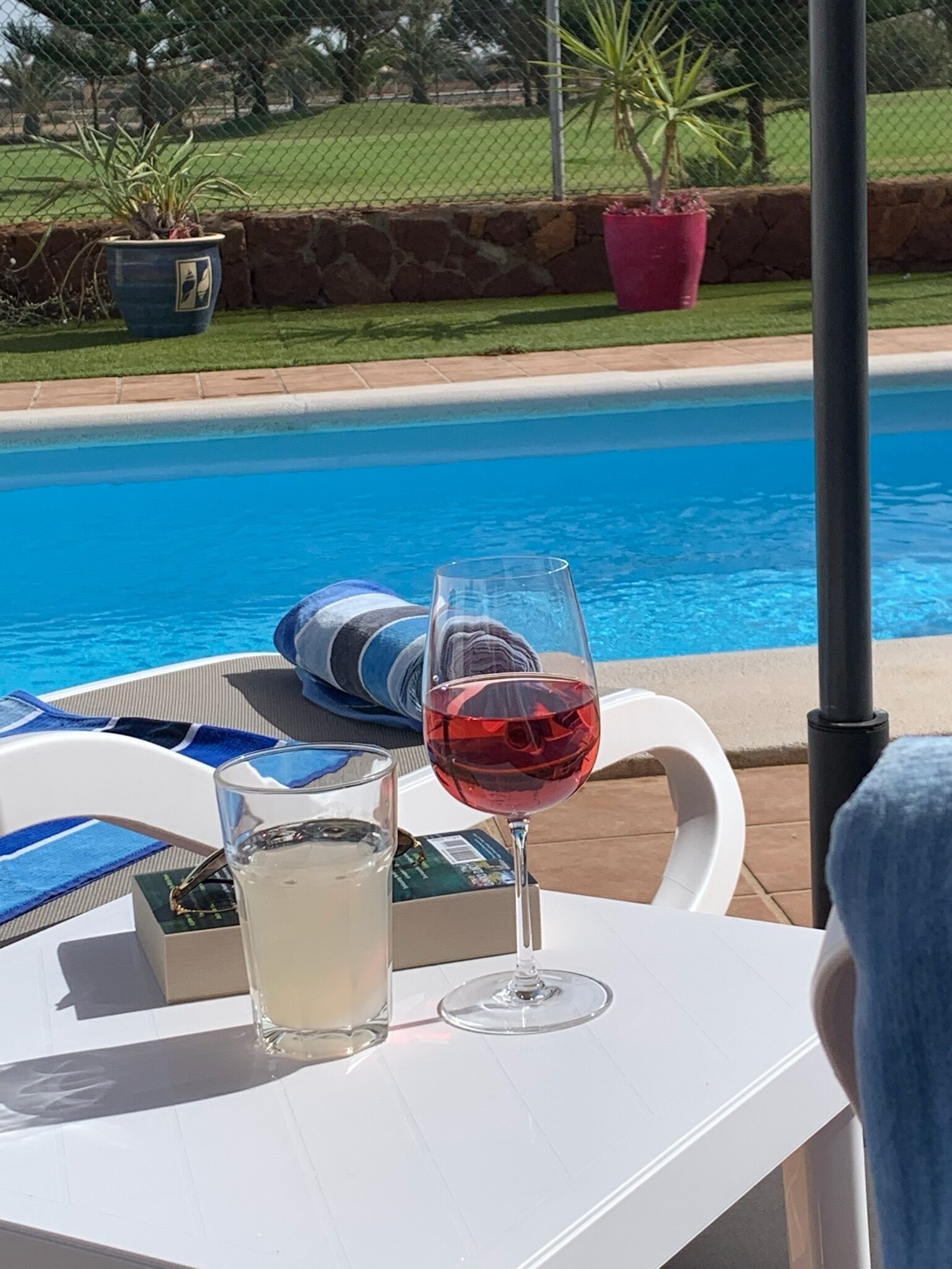 Une table au bord de la piscine, avec un verre de vin rouge et un verre de vin blanc, une serviette et un livre.