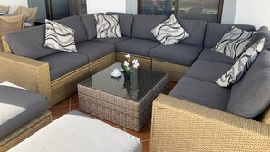 Un sofá modular con cojines grises y almohadas estampadas, una mesita de centro de mimbre con una taza blanca y flores, y vistas al exterior a través de ventanas amplias.