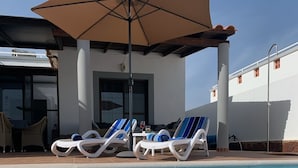 Un área de piscina con sillones de descanso y una sombrilla, un edificio de fachada blanca y un cielo azul despejado.