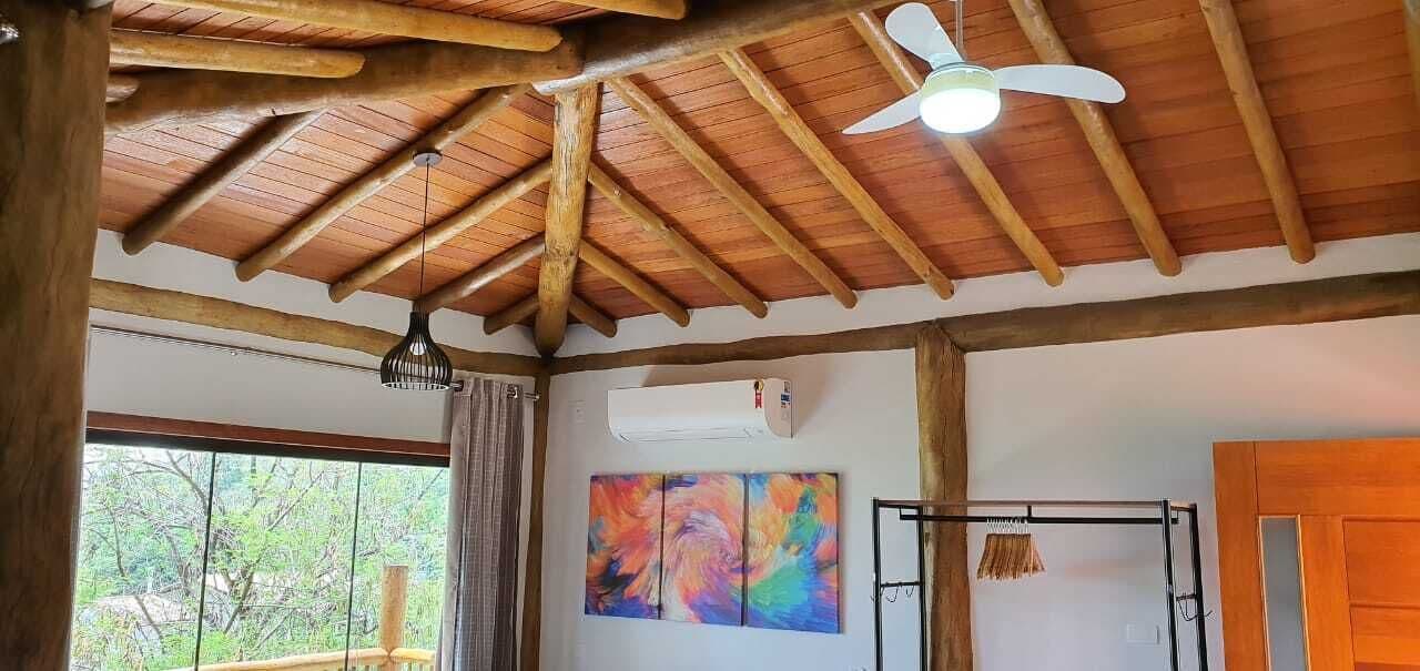 A room with a wooden ceiling, a ceiling fan, a window with a view of trees, an air conditioning unit, and a colorful abstract painting.