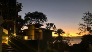A building with a balcony overlooking a sunset, a staircase leading up to it, and a view of the water in the distance. - Siriúba Reserve (Ilhabela)