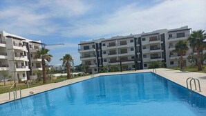 A rectangular swimming pool with a clear blue water surface, surrounded by a tiled deck. In the background, there are multi-story residential buildings with balconies and a clear sky. - Quiet spot in Cabo negro (Cabo Negro)