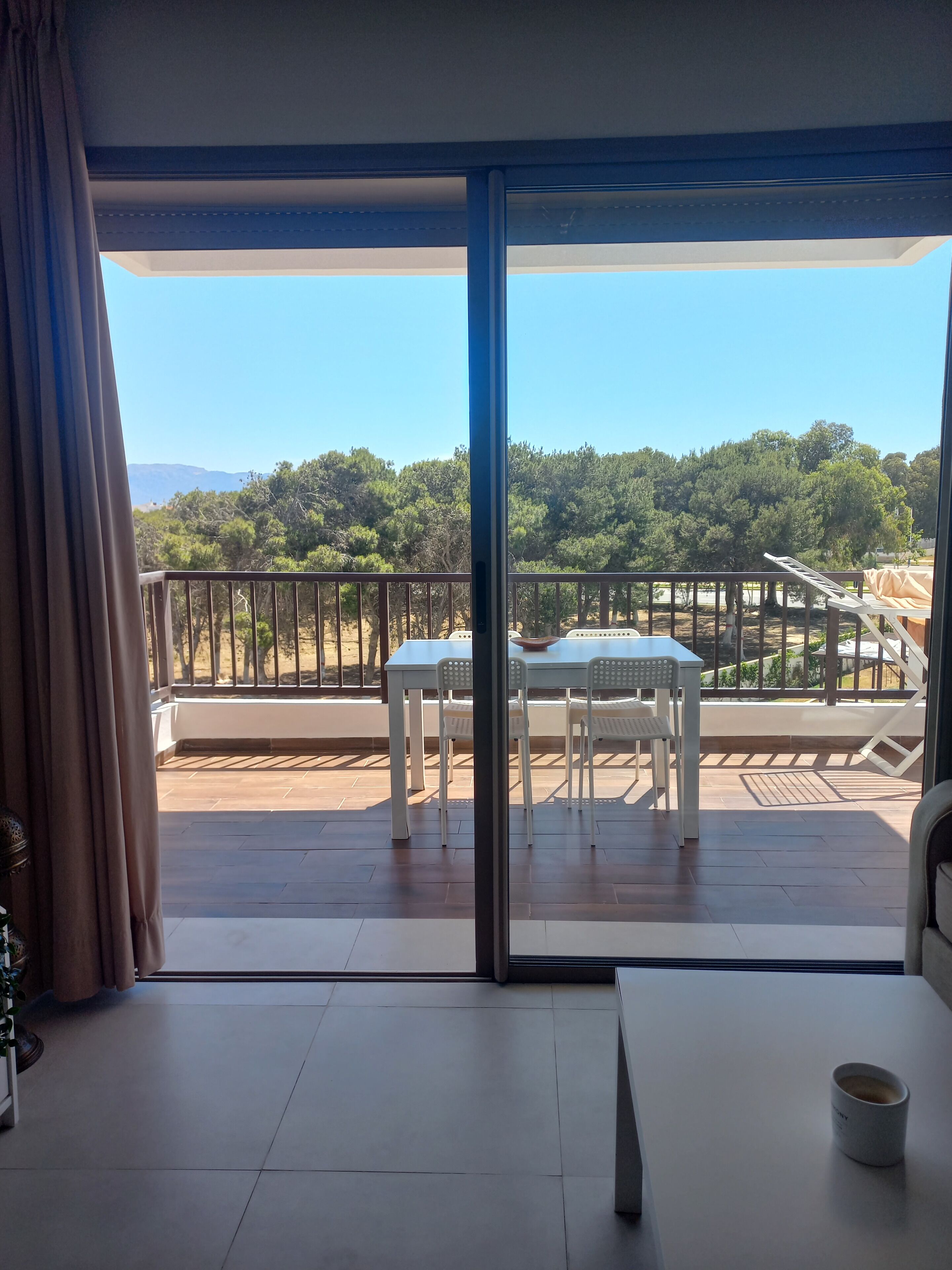 Un balcon avec une table et des chaises, une vue sur les arbres et les collines, et un ciel dégagé.