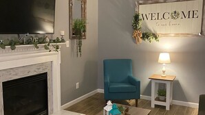 Een woonkamer met een open haard, een televisie, een blauwgroene fauteuil, een kleintje tafeltje met een plant, en een welkomstbord.