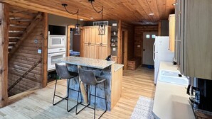 Interior rústico de cabana com escada de madeira, cozinha com ilha central, banquetas de bar e mesa de jantar.