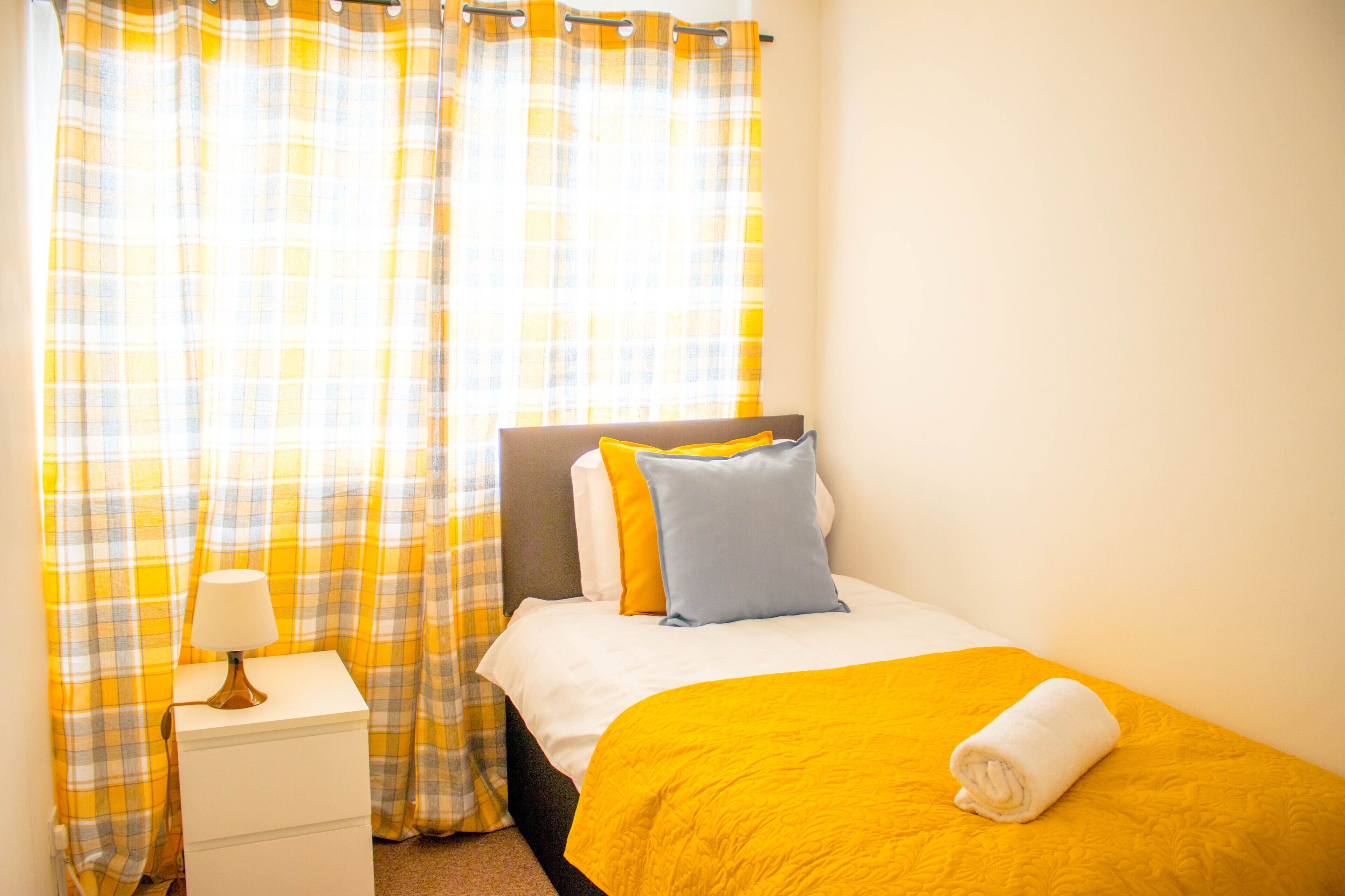 A neatly made bed with a yellow bedspread and pillows, a white bedside table with a lamp, and plaid curtains.