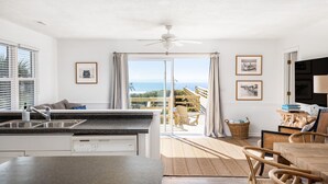 A modern kitchen with a large island, a dining area with wicker furniture, and a balcony with a view of the ocean.