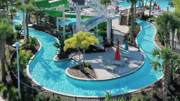 A pool area with a water slide, a small bridge, and a circular pool surrounded by palm trees and lounge chairs. - Family Fun Oasis|Arcade|Game Room|FREE Pool Heat (Davenport)