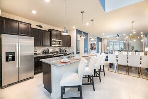 Uma cozinha moderna com armários escuros, eletrodomésticos de aço inoxidável, uma ilha central com banquetas brancas e iluminação pendente.