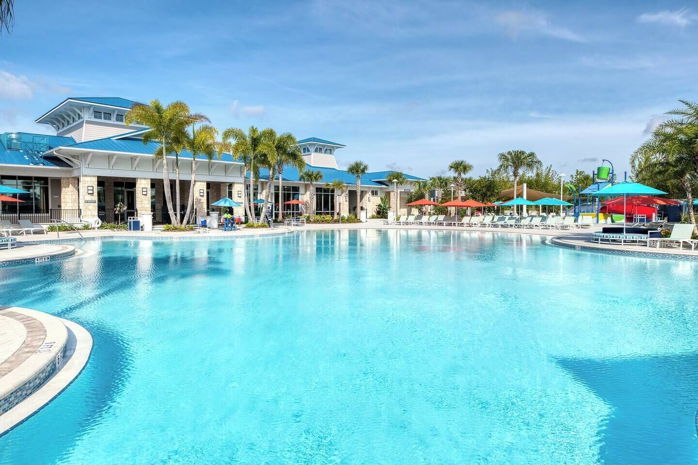 A large swimming pool with lounge chairs and umbrellas, surrounded by buildings with blue roofs and palm trees.