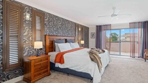 A bedroom with a large bed, a nightstand, a wooden dresser, a ceiling fan, and a view of the outdoors through a sliding glass door. - Riverside Breeze - private luxury family retreat with pool (Port Macquarie)