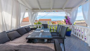 Un espacio de estar al aire libre con techo, con mesa y sillas, que ofrece vistas a un paisaje urbano con edificios de tejados rojos y vista al mar.