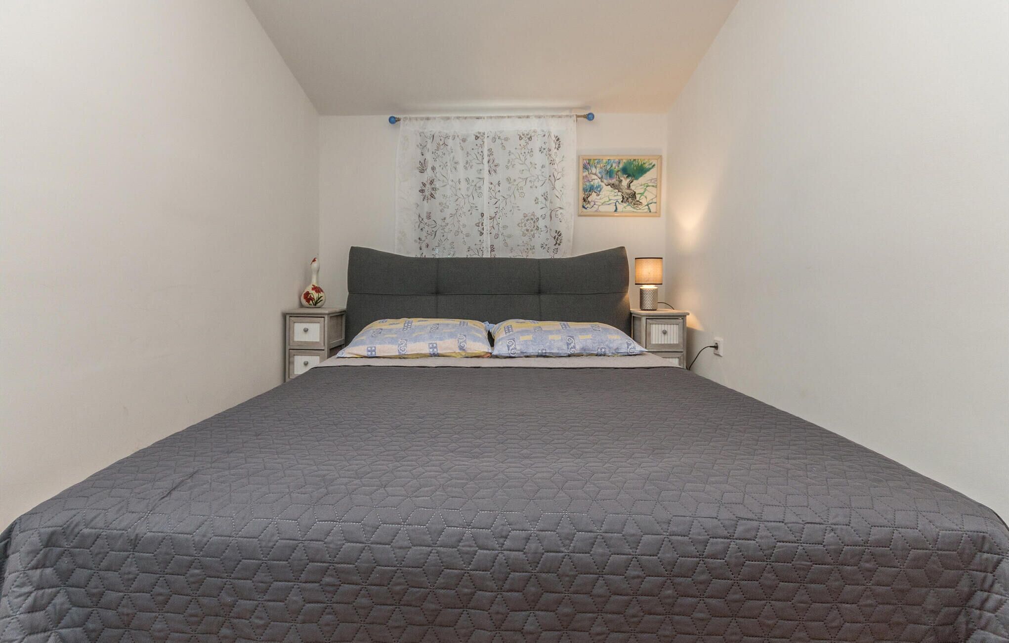 A bedroom with a bed, two bedside tables, a nightstand, a lamp, a floral curtain, and a painting on the wall.