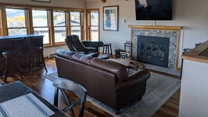 A spacious living room with a leather sofa, a fireplace, a television, and large windows offering a view of the outdoors. - Spacious In-Town Condo | Pool | Hot Tub (Durango)