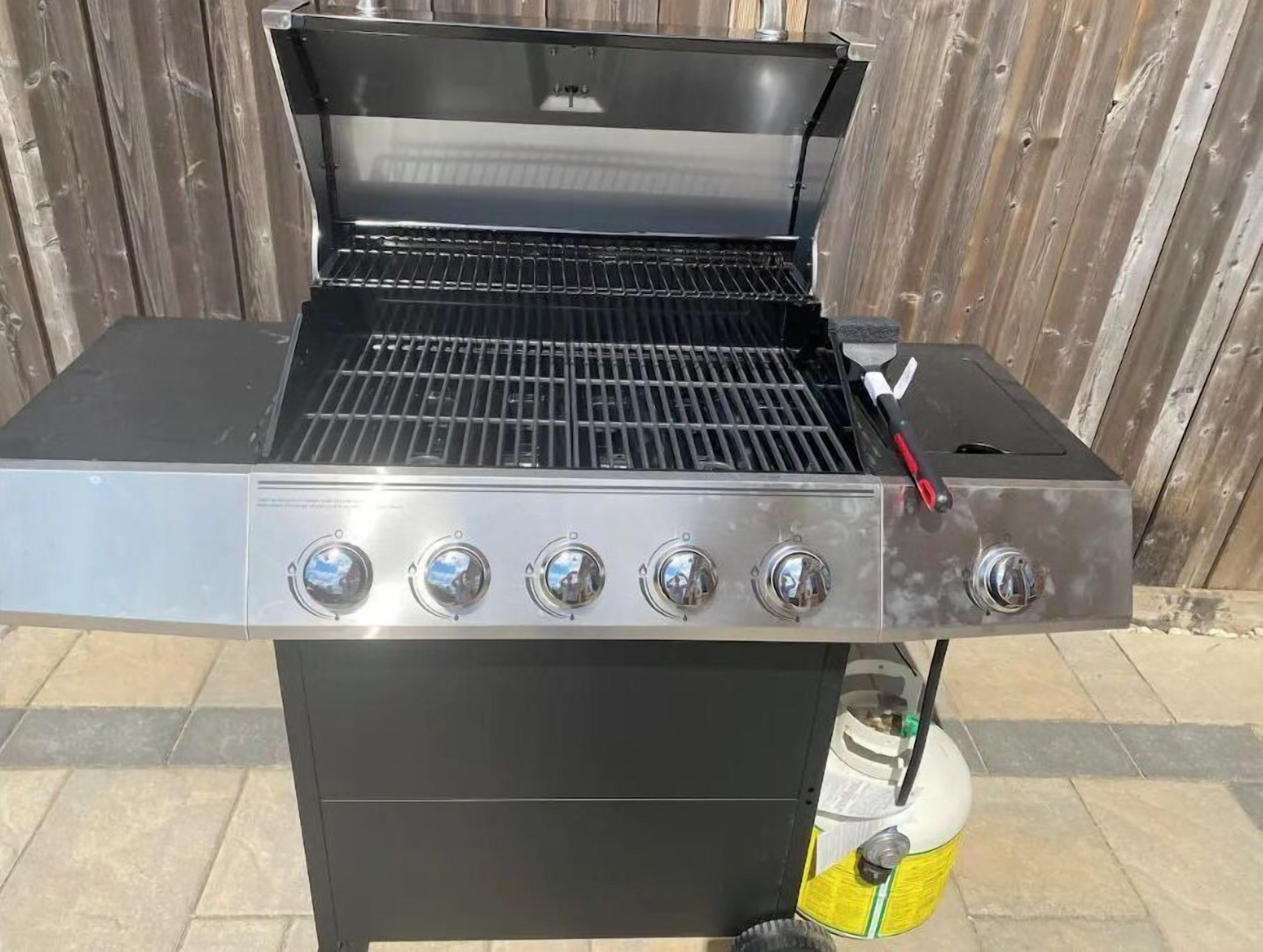 A stainless steel barbecue grill with a gas tank and a red-handled tool.
