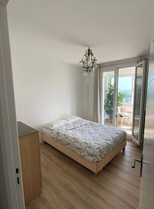 Un dormitorio pequeño con cama, una mesita de noche de madera, una araña y un ventanal corredero que da a un balcón con una silla y una mesa.